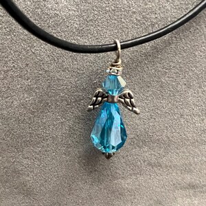 Op de afbeelding: Een delicate ketting met een blauwe kristallen engel hanger. De engel heeft zilveren vleugels en een kristallen lichaam. De ketting is aan een zwart koord.