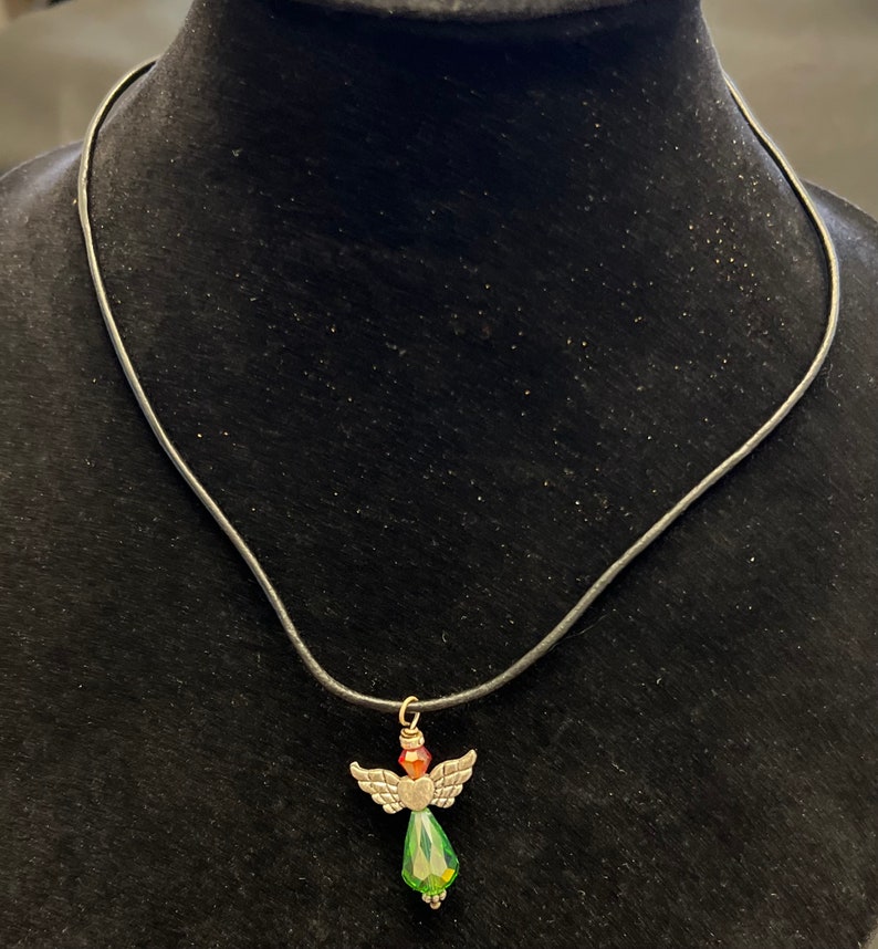 Op de afbeelding: Een zilveren engel hanger ketting met een groene en rode kristallen hanger. De ketting is aan een zwart lederen koord.