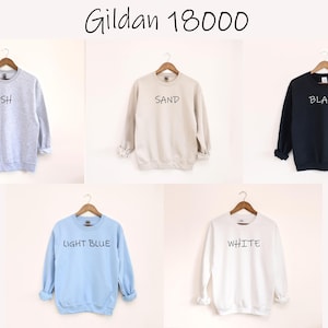 May include: Five Gildan 18000 sweatshirts in various colors: ash, sand, black, light blue, and white. Each sweatshirt is displayed on a wooden hanger against a neutral background. The color name is printed in black on each sweatshirt.