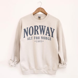 以下が含まれることがあります： 胸に紺色の「NORWAY」の文字がアーチ状に描かれたベージュのスウェットシャツ。「NORWAY」の下には「ALT FOR NORGE」と年号「1905」の文字。スウェットシャツは木製のハンガーにかかっています。
