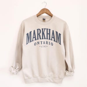 May include: A beige crewneck sweatshirt with the word "MARKHAM" arched across the chest in navy blue lettering, with "ONTARIO" and "*1793*" below. The sweatshirt is hanging on a wooden hanger.