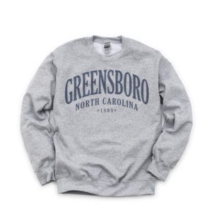 May include: A heather gray sweatshirt with the text "GREENSBORO NORTH CAROLINA *1808*" in navy blue arched across the chest. The sweatshirt has a crew neck and long sleeves.