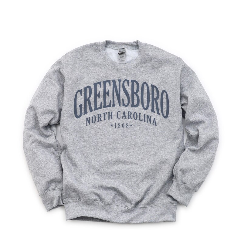 May include: A heather gray sweatshirt with the text "GREENSBORO NORTH CAROLINA *1808*" in navy blue arched across the chest. The sweatshirt has a crew neck and long sleeves.
