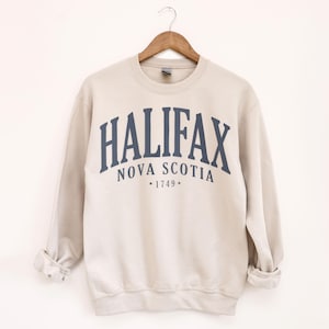Può includere: Una felpa beige con la scritta "HALIFAX" in blu navy ad arco sul petto, con "NOVA SCOTIA" e "*1749*" sotto. La felpa è appesa a una gruccia di legno.