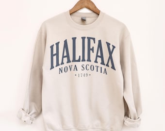 May include: A beige sweatshirt with the word "HALIFAX" in navy blue arched across the chest, with "NOVA SCOTIA" and "*1749*" below. The sweatshirt is hanging on a wooden hanger.