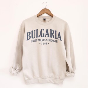 May include: A beige sweatshirt with the word "BULGARIA" in navy blue arched across the chest. Below the word is the phrase "UNITY MAKES STRENGTH" and the year "1908". The sweatshirt is hanging on a wooden hanger.