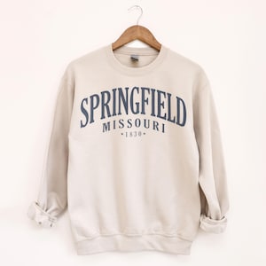 Puede incluir: Sudadera beige de cuello redondo con el texto "SPRINGFIELD MISSOURI *1830*" en azul marino arqueado en el pecho. La sudadera está colgada de una percha de madera sobre un fondo blanco.