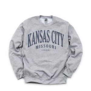 May include: A grey sweatshirt featuring the navy blue text "KANSAS CITY MISSOURI *1850*" in an arched design. The sweatshirt has a crew neck and long sleeves.