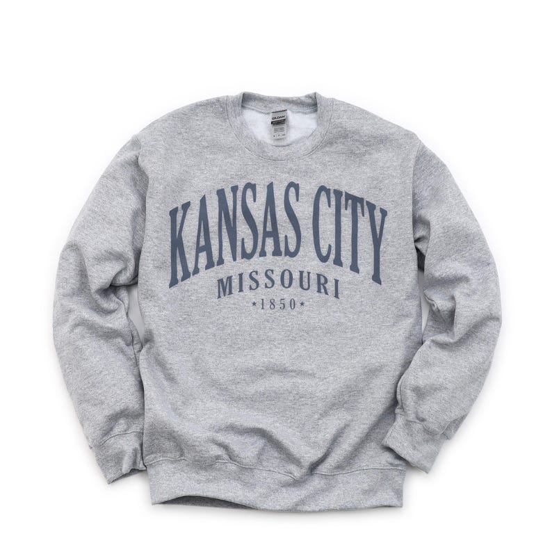 May include: A grey sweatshirt featuring the navy blue text "KANSAS CITY MISSOURI *1850*" in an arched design. The sweatshirt has a crew neck and long sleeves.
