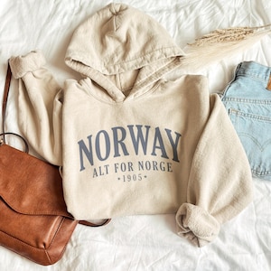 May include: A beige hooded sweatshirt featuring the dark blue text "NORWAY ALT FOR NORGE *1905*" printed on the front. The garment has long sleeves with ribbed cuffs, a drawstring hood, and a front kangaroo pocket, presented in a casual style.