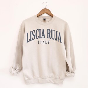 May include: A beige sweatshirt with the text "LISCIA RUJA ITALY" in navy blue arched across the chest. The sweatshirt is hanging on a wooden hanger against a white background. The sleeves are cuffed.