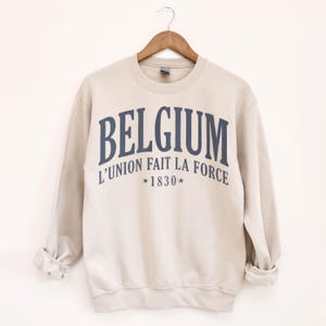 May include: A beige sweatshirt with the word "BELGIUM" in navy blue arched text. Below, the text "L'UNION FAIT LA FORCE" and "*1830*" are printed. The sweatshirt is hanging on a wooden hanger.