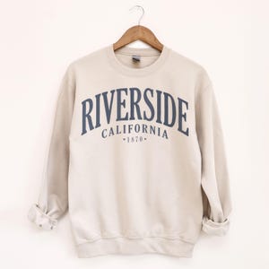 May include: A beige sweatshirt with the word "RIVERSIDE" arched across the chest in navy blue lettering. Below, the word "CALIFORNIA" and the year "1870" are printed in a smaller font. The sweatshirt is hanging on a wooden hanger.