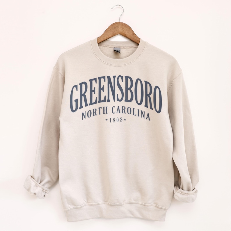 May include: A beige sweatshirt with the word "GREENSBORO" arched across the chest in navy blue lettering. Below the city name are the words "NORTH CAROLINA" and the year "1808". The sweatshirt is hanging on a wooden hanger.
