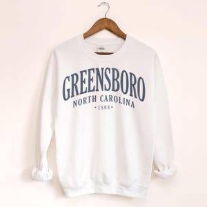 May include: White sweatshirt with the word "GREENSBORO" in navy blue arched across the chest. Below the word "GREENSBORO" is "NORTH CAROLINA" and "*1808*". The sweatshirt is hanging on a wooden hanger.