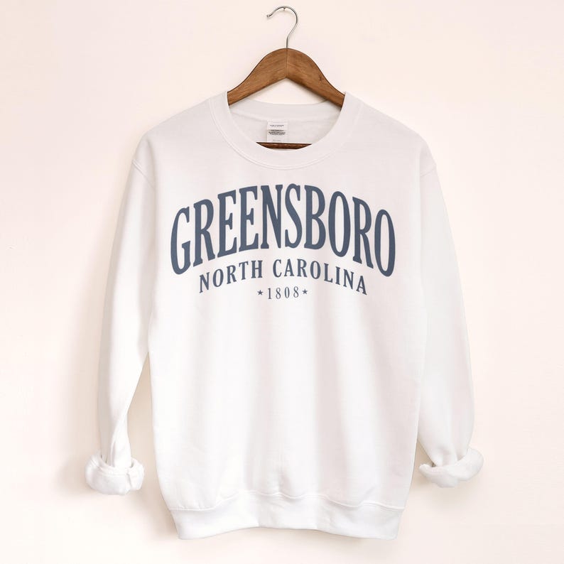 May include: White sweatshirt with the word "GREENSBORO" in navy blue arched across the chest. Below the word "GREENSBORO" is "NORTH CAROLINA" and "*1808*". The sweatshirt is hanging on a wooden hanger.