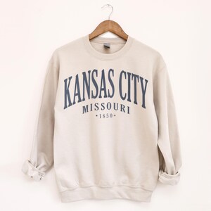 May include: A beige sweatshirt featuring the navy blue text "KANSAS CITY MISSOURI *1850*" arched across the chest. The garment is suspended on a wooden hanger against a white backdrop. The sleeves are rolled up.