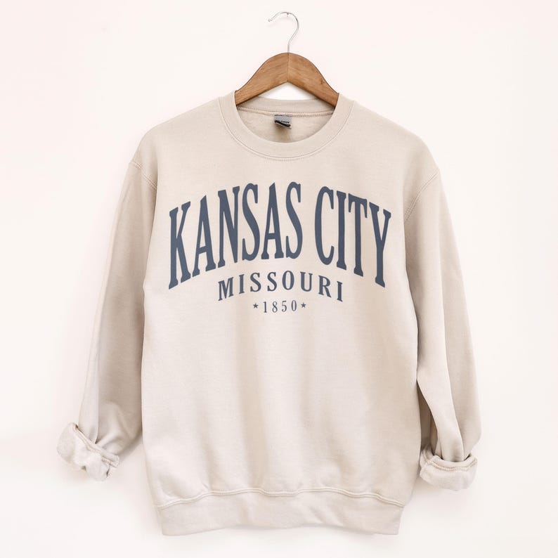 May include: A beige sweatshirt featuring the navy blue text "KANSAS CITY MISSOURI *1850*" arched across the chest. The garment is suspended on a wooden hanger against a white backdrop. The sleeves are rolled up.