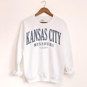 May include: White sweatshirt with navy blue text reading "KANSAS CITY MISSOURI *1850*". The garment is suspended from a wooden hanger.