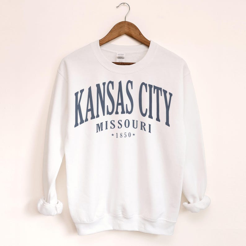 May include: White sweatshirt with navy blue text reading "KANSAS CITY MISSOURI *1850*". The garment is suspended from a wooden hanger.