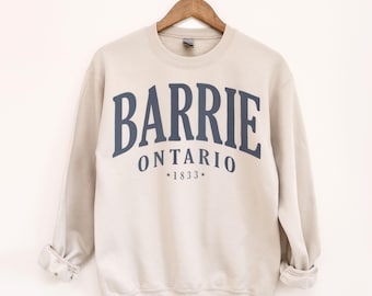 May include: A beige crewneck sweatshirt with the word "BARRIE" in navy blue arched across the chest, with "ONTARIO" and "*1833*" below. The sweatshirt is hanging on a wooden hanger.