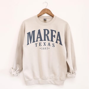 May include: A beige sweatshirt with the word "MARFA" arched across the chest in navy blue letters, with "TEXAS" and "*1883*" below. The sweatshirt is hanging on a wooden hanger.