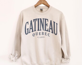 May include: A beige crewneck sweatshirt with the word "GATINEAU" arched across the chest in navy blue lettering. Below the word "GATINEAU" is the word "QUEBEC" and the year "1800". The sweatshirt is hanging on a wooden hanger.