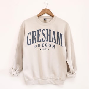 Può includere: Felpa beige con la scritta "Gresham" in blu navy ad arco sul petto, con "Oregon" e "1905" sotto. La felpa è appesa a una gruccia di legno.