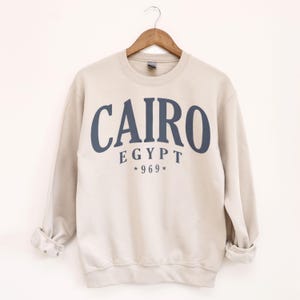 May include: A beige sweatshirt with the word "CAIRO" in navy blue block letters. Below the word "CAIRO" is the word "EGYPT" and the numbers "*969*". The sweatshirt is hanging on a wooden hanger.
