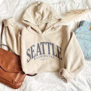 以下が含まれることがあります： 胸元にアーチ状のフォントでSEATTLE WASHINGTON 1851とダークブルーの文字がプリントされたベージュのフード付きスウェットシャツ。長袖のプルオーバーには、ドローストリング付きのフード、リブ編みの袖口、フロントにカンガルーポケットが付いています。