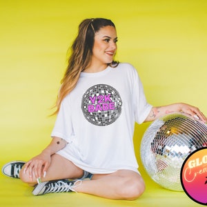 May include: A white t-shirt featuring a pink and black disco ball graphic with the text "Y2K BABE". The person wearing the shirt is sitting on a yellow background with a silver disco ball.