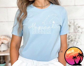 Könnte beinhalten: Hellblaues T-Shirt mit dem Schriftzug "Magical Runner" in weißer Schrift und Blockbuchstaben. Ein weißes Stern- und Wirbeldesign befindet sich über dem Text. Das T-Shirt hat kurze Ärmel und einen Rundhalsausschnitt.