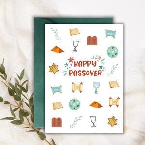 May include: A white greeting card with a green envelope, featuring colorful illustrations and the text "HAPPY PASSOVER." The card includes images of matzah, a Star of David, a chalice, and other Passover symbols. The background is a soft, white fabric.