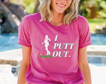 May include: Berry-colored V-neck t-shirt with the text "I Putt Out." A white silhouette of a golfer is on the left side of the text. The golfer is standing on a patch of green grass. The shirt is a casual style.