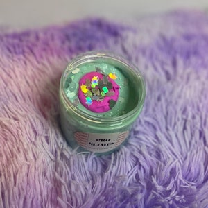 May include: A clear plastic jar filled with light green slime with a pink and iridescent glitter swirl on top. The jar has a label that reads "PRO SLIMES" and "Unicorn cake batter".