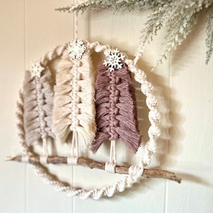 May include: A macrame wall hanging with three beige and pink trees with wooden snowflake ornaments. The trees are attached to a branch and the hanging is made with a white rope.