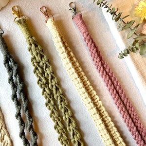 Peut inclure: Gros plan sur cinq lani&egrave;res macram&eacute; de couleurs vari&eacute;es : beige, gris, vert olive, cr&egrave;me et mauve. Chaque lani&egrave;re est dot&eacute;e d'un mousqueton en m&eacute;tal. Les lani&egrave;res sont dispos&eacute;es en diagonale sur une surface blanche, avec des fleurs et de la verdure en arri&egrave;re-plan.