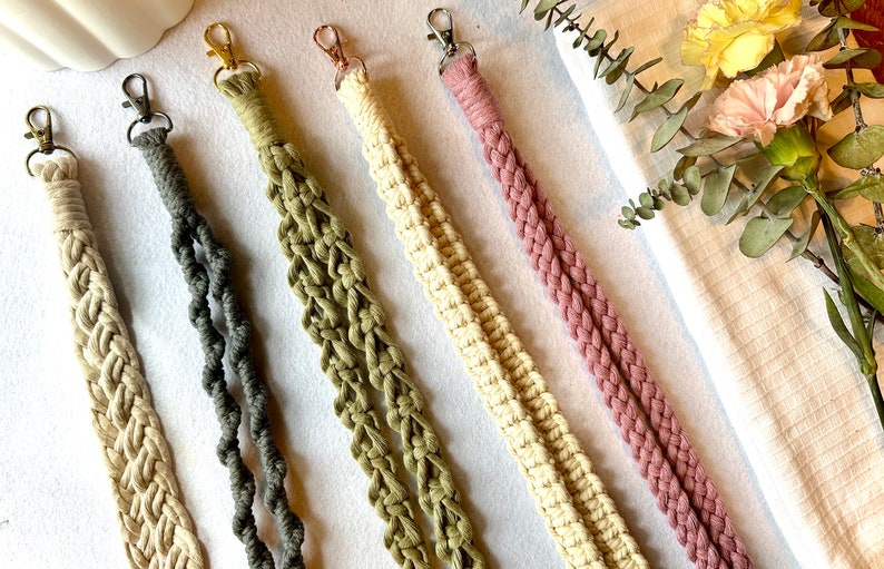 Peut inclure: Gros plan sur cinq lani&egrave;res macram&eacute; de couleurs vari&eacute;es : beige, gris, vert olive, cr&egrave;me et mauve. Chaque lani&egrave;re est dot&eacute;e d'un mousqueton en m&eacute;tal. Les lani&egrave;res sont dispos&eacute;es en diagonale sur une surface blanche, avec des fleurs et de la verdure en arri&egrave;re-plan.