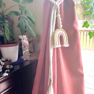 May include: A white twisted rope curtain tieback featuring a decorative macrame rainbow in shades of mustard yellow and dusty pink, accented with four natural wooden beads and finished with two long rope tassels at the bottom ends.