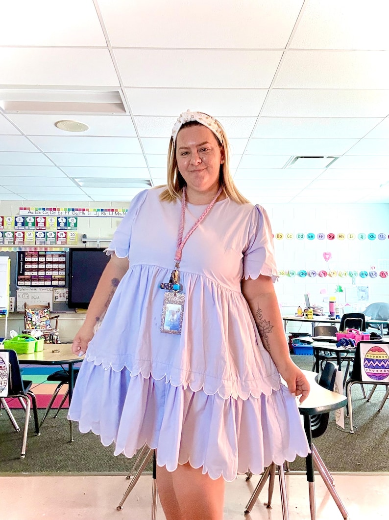 Peut inclure: Robe babydoll violet clair avec ourlet festonn&eacute; et manches courtes. La robe est port&eacute;e avec un bandeau blanc. Un cordon rose avec un porte-nom est autour du cou. Le fond est une salle de classe.