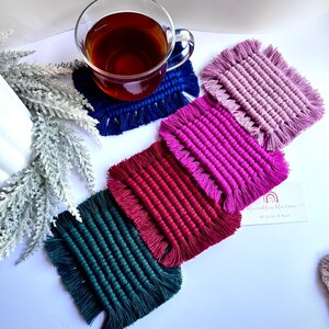 May include: Five square crocheted coasters with fringe in shades of teal, burgundy, magenta, and lavender. The coasters are arranged in a stack with the teal coaster on the bottom and the lavender coaster on top. The coasters are handmade and have a unique, textured design.