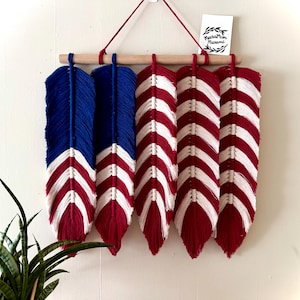 May include: Macrame feather wall hanging in red, white, and blue. The feathers are attached to a wooden dowel and suspended by a red cord. The design resembles the American flag. A small white tag with text is visible.