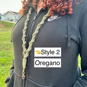 Peut inclure: Un cordon de macram&eacute; vert avec un fermoir dor&eacute;. Le cordon est &eacute;tiquet&eacute; "Style 2 Oregano" dans un encadr&eacute; blanc. La personne sur l'image porte un sweat &agrave; capuche noir.