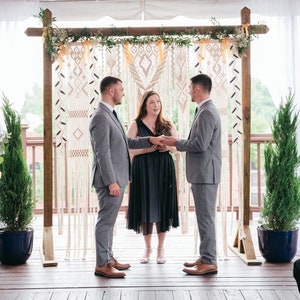May include: Two people in suits stand facing each other, holding hands during a wedding ceremony. A woman in a black dress stands between them, officiating the ceremony. A macrame backdrop hangs behind them.