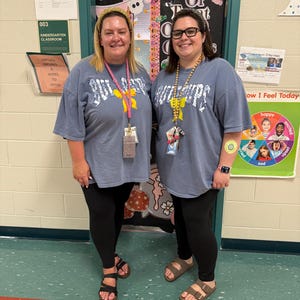 Peut inclure: Deux femmes portant des t-shirts bleus assortis avec le mot "Bulldogs" imprim&eacute; dessus. Elles se tiennent dans une salle de classe, avec une porte d&eacute;cor&eacute;e pour Halloween en arri&egrave;re-plan. Elles portent des leggings noirs et des sandales.