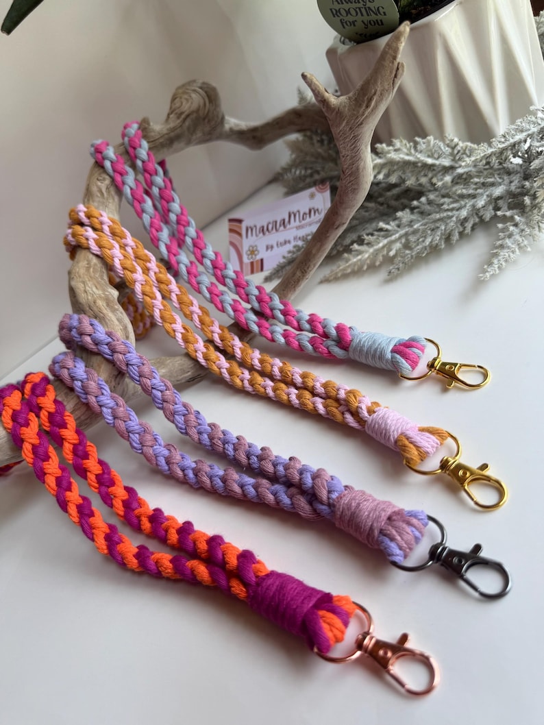 Peut inclure: Quatre lani&egrave;res macram&eacute; aux tons de rose, orange, violet et beige. Chaque lani&egrave;re est tress&eacute;e avec une combinaison de couleurs diff&eacute;rente et comporte un fermoir en m&eacute;tal. Les lani&egrave;res sont pr&eacute;sent&eacute;es sur une surface blanche.