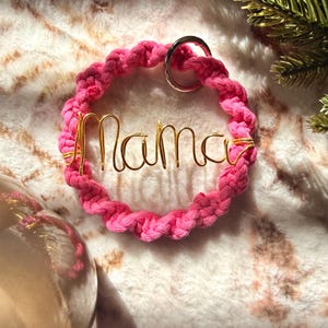 May include: A pink macrame keychain with the word "mama" in gold wire. The keychain has a silver ring and is set against a soft, textured background. The design is simple and elegant.