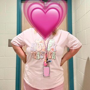 Peut inclure: Un t-shirt rose clair avec l'inscription "Happy Teacher" dans une police r&eacute;tro. Le t-shirt est associ&eacute; &agrave; un pantalon &agrave; carreaux roses et blancs. Un cordon avec un porte-nom et des breloques color&eacute;es est autour du cou.