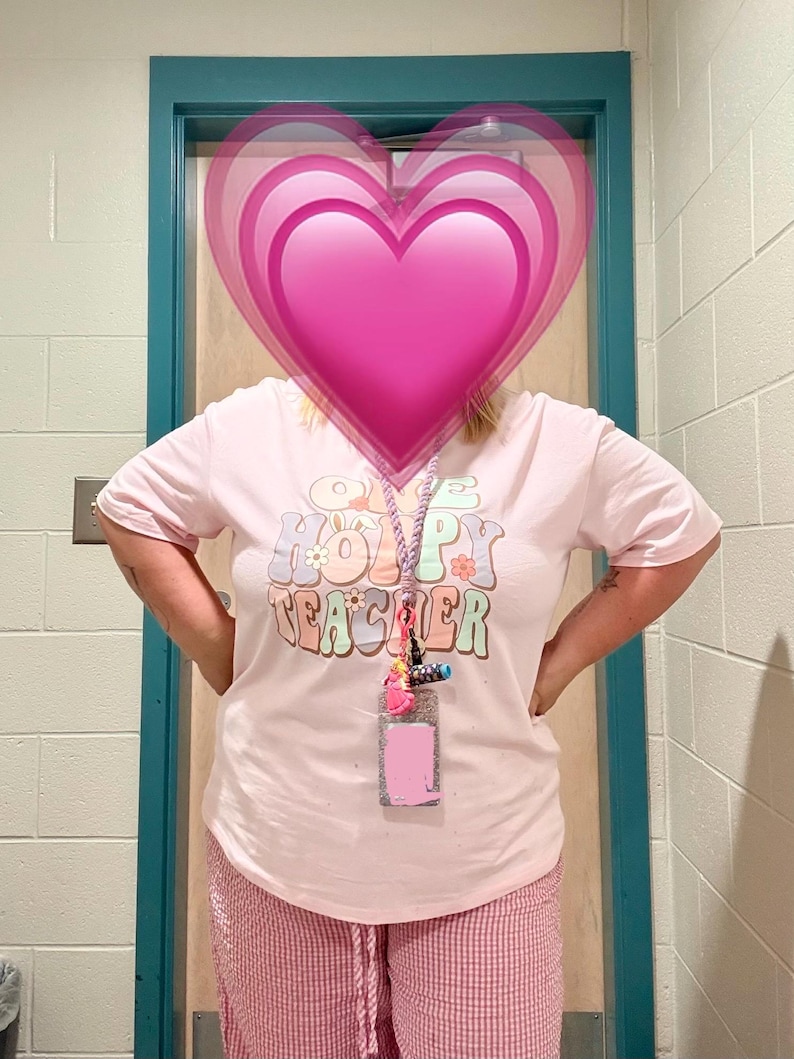 Peut inclure: Un t-shirt rose clair avec l'inscription "Happy Teacher" dans une police r&eacute;tro. Le t-shirt est associ&eacute; &agrave; un pantalon &agrave; carreaux roses et blancs. Un cordon avec un porte-nom et des breloques color&eacute;es est autour du cou.