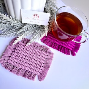 May include: Two square macrame coasters with fringe. The coaster on the left is a light purple color and the coaster on the right is a bright pink color. A glass of tea sits on the pink coaster.
